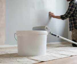 A person touches a freshly painted light grey wall. A white paint bucket and roller sit on a drop cloth, showing home improvement.