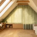 Bright, modern attic room with sloped ceiling. Custom sage green curtains follow the incline, filtering light. Cozy armchair, window seat, plants.