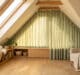 Bright, modern attic room with sloped ceiling. Custom sage green curtains follow the incline, filtering light. Cozy armchair, window seat, plants.