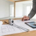 Person planning construction with blueprints, a tape measure, and a tablet calculator on a wooden desk in a bright, unfinished room.