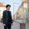 Gender-neutral individual thoughtfully observes a transparent digital map of Dijon, blurred historic buildings in background.