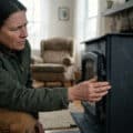 A person in earthy tones inspects a wood stove, touching a seam with a skeptical expression, evaluating its manufacturing quality in a cozy home.
