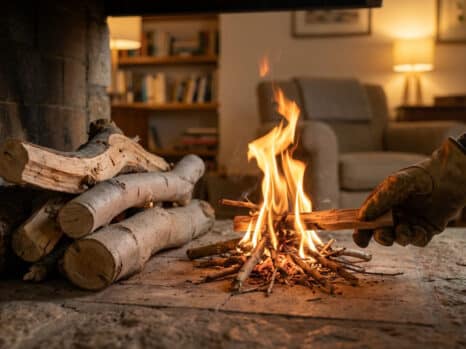 Close-up of a hand in work gloves adding small fig wood to a quick-burning fire in a rustic fireplace, with more logs nearby and a cozy living room in the background.