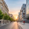 Sunset street scene in a French city. Haussmannian buildings left, modern right. People walk on clean sidewalks under a warm sky.