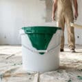 White bucket with vibrant emerald green paint dripping, on a drop cloth. A painter with roller stands in a spacious, bright renovated room.