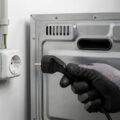 Gloved hand connecting an electric oven's power plug to a wall socket with visible wiring, emphasizing safe electrical setup.