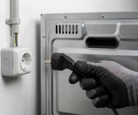 Gloved hand connecting an electric oven's power plug to a wall socket with visible wiring, emphasizing safe electrical setup.