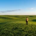 Photorealistic wide-angle shot of a person standing in a vast, vibrant green landscape under a clear blue sky, contemplating the expansive hills.