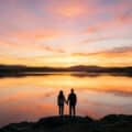 Un couple se tient la main devant un lac au coucher du soleil avec un ciel aux teintes orangées.