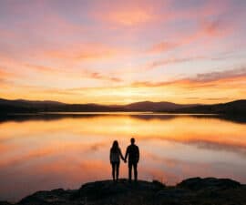 Un couple se tient la main devant un lac au coucher du soleil avec un ciel aux teintes orangées.