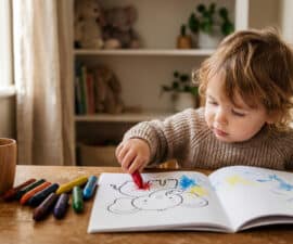 Un jeune enfant blond colorie avec un crayon rouge un dessin d'éléphant dans un cahier posé sur une table en bois.
