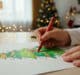 Main d'un enfant utilisant un crayon rouge pour colorier un dessin de sapin de Noël sur une table décorée.