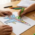 Un enfant colorie un dessin d'avion de chasse avec des crayons de couleur sur une table en bois.