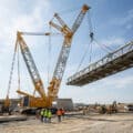 Une immense grue sur chenilles déplace une structure métallique de pont sous un ciel bleu sur un chantier de construction.