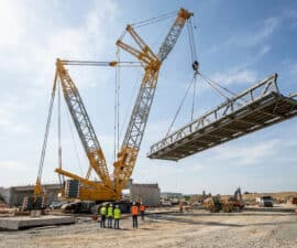 Une immense grue sur chenilles déplace une structure métallique de pont sous un ciel bleu sur un chantier de construction.