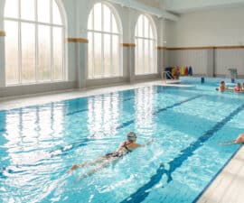 Piscine intérieure lumineuse avec des nageurs, une personne qui nage et un homme assis au bord du bassin.