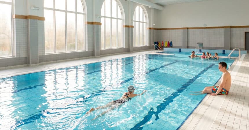 Piscine intérieure lumineuse avec des nageurs, une personne qui nage et un homme assis au bord du bassin.